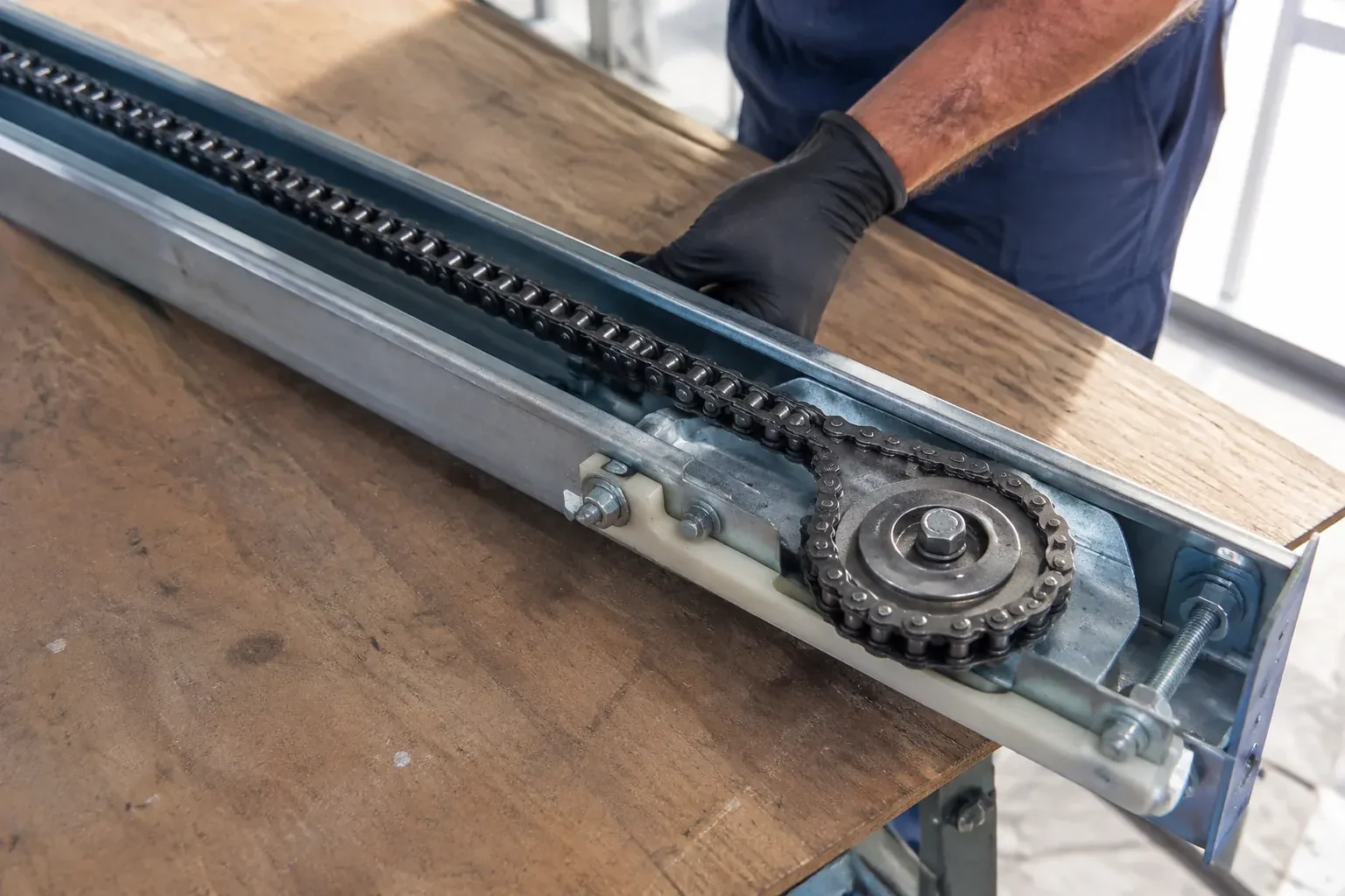 Gloved hand resting on a steel opener rail laid on a wooden bench, showing a drive chain looped around a sprocket wheel. — Chain-Drive Opener Repair, Silverthorne