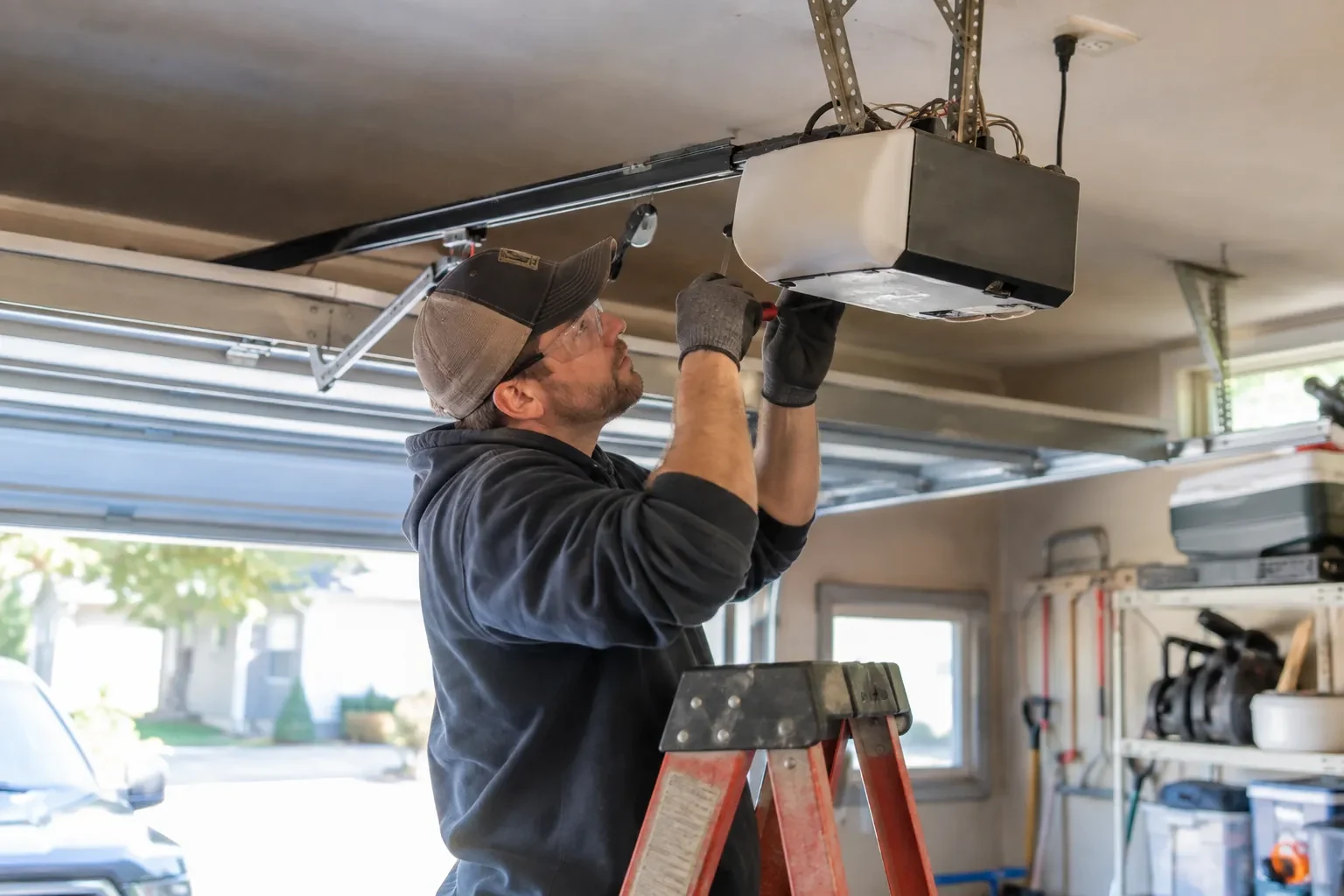 Technician in safety glasses on an orange ladder, holding a small screwdriver against a white and black opener with wires showing.